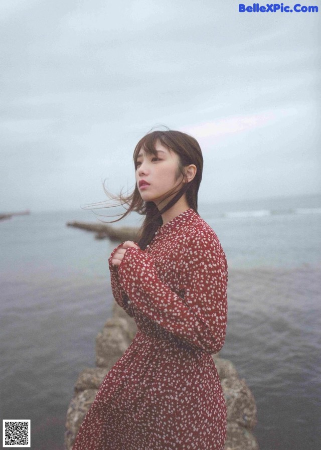 A woman in a red dress standing on a rock by the ocean.